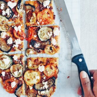 Flatbread with roasted vegetables and feta, sliced with hand holding a knife