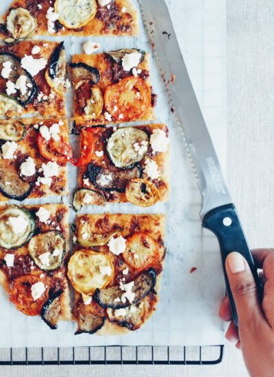 Flatbread with roasted vegetables and feta, sliced with hand holding a knife