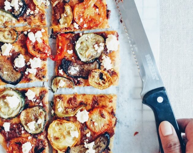 Flatbread with roasted vegetables and feta, sliced with hand holding a knife