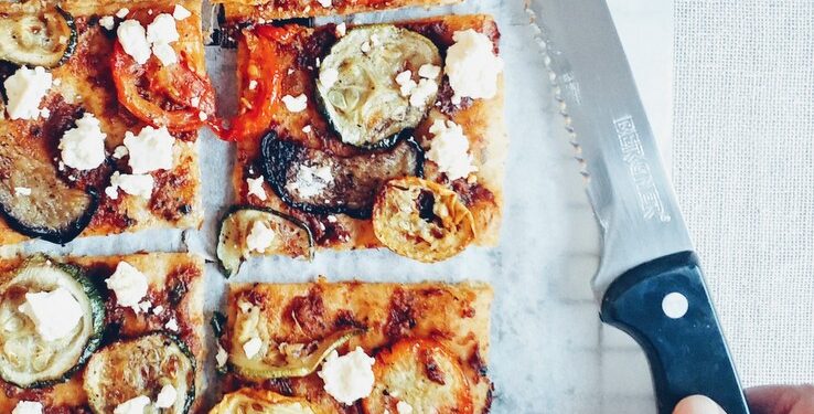 Flatbread with roasted vegetables and feta, sliced with hand holding a knife