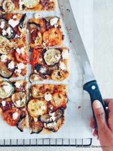 Flatbread with roasted vegetables and feta, sliced with hand holding a knife